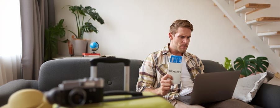 Angry man holding his flight tickets, staring upset at laptop screen, having problems with his holiday tour.