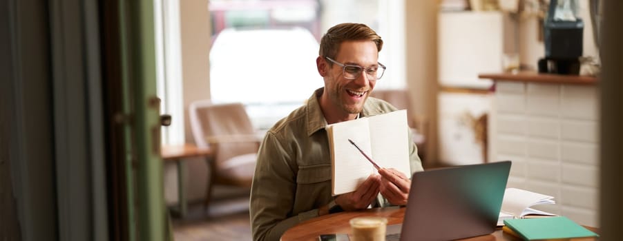 Portrait of young handsome man in glasses, private tutor teaching student online, pointing at his notebook, showing something, video chats via laptop application, working remotely in cafe.