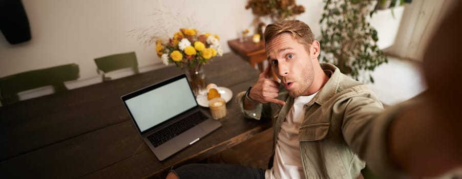 Handsome stylish young man takes selfie in cafe while working or studying remotely with laptop, shows call phone hand sign and smiling at mobile phone camera.