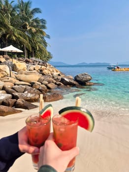 Two hands clinking glasses of watermelon mojito with fruit garnish against stunning Nha Trang beach backdrop. Turquoise sea, white sand, sunny Vietnam paradise