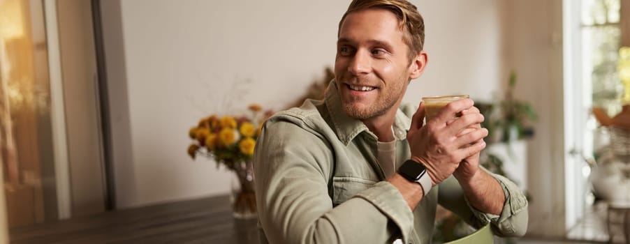 Cafe, leisure and lifestyle concept. Portrait of handsome young man sits on chair with glass of coffee, drinks his beverage and smiling.