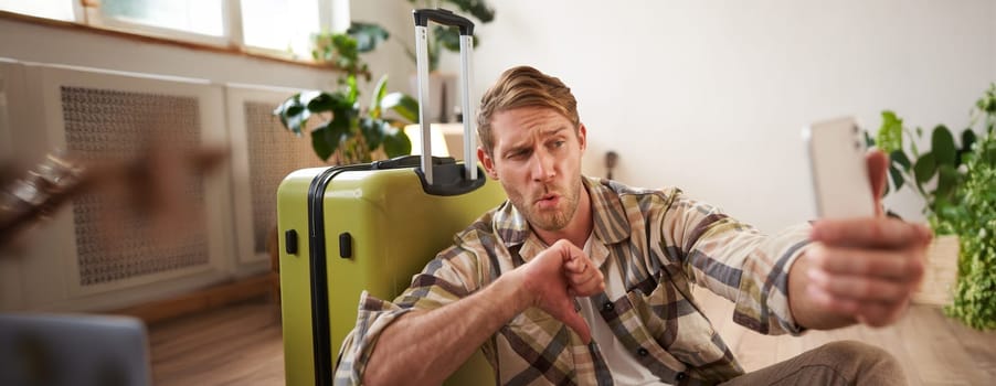 Disappointed tourist, young man giving negative feedback, shows thumbs down, takes photo on smartphone, sits near travel suitcase.