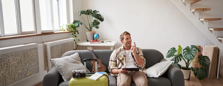 Portrait of young man in living room, sitting with tablet in hands, has suitcase packed and two plane tickets, preparing to go abroad, going on holiday.