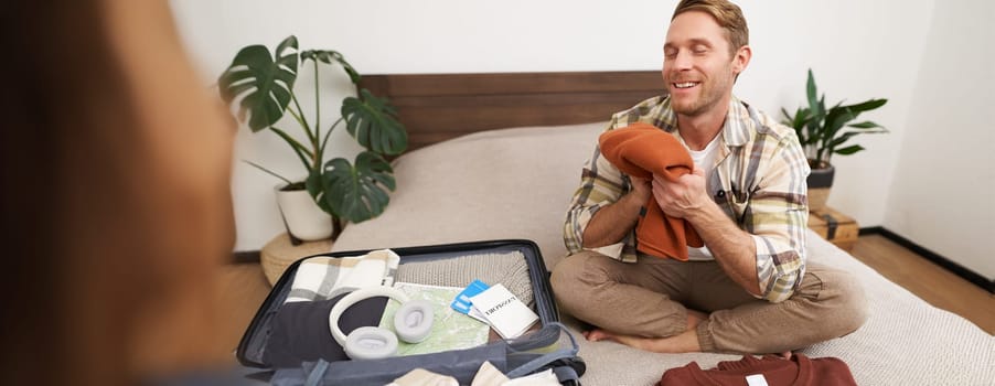 Portrait of smiling young man sits on bed with suitcase, packing for summer holiday, smelling clean and fresh clothes, goes on vacation.