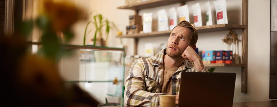 Man looking concentrated while working remotely from a coffee shop. Business owner sitting in cafe with laptop, drinking cappuccino, looking aside with thoughtful face, getting inspiration.