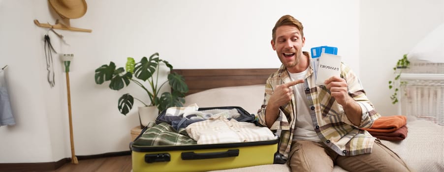 Portrait of handsome young man, traveller shows his passport and two tickets, packing suitcase with clothes, going on vacation. Tourism and holiday concept