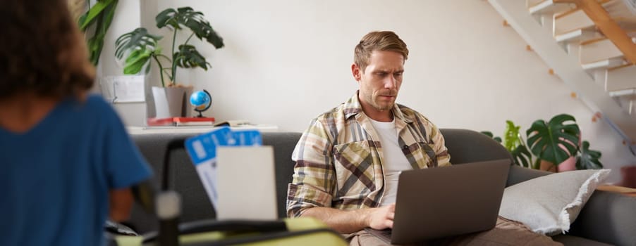 Portrait of serious and concentrated man, sits with laptop in living room, has two plane tickets, plans holiday, goes online.