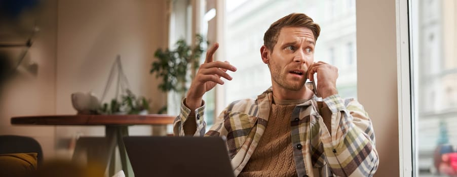 Portrait of serious businessman talking over the phone, sitting in chair in a cafe with laptop, working on project, discussing work-related issues on telephone, looks behind him.