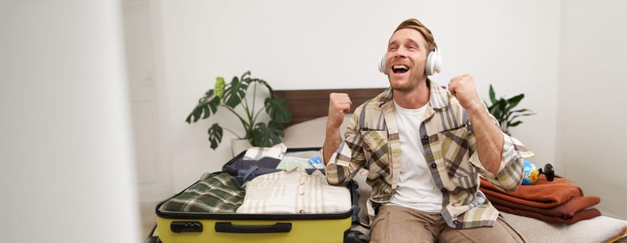 Excited young man in headphones, gets ready for vacation, goes on holiday, packs things in suitcase, listens to music in earphones, sits near his luggage. Tourism and lifestyle concept