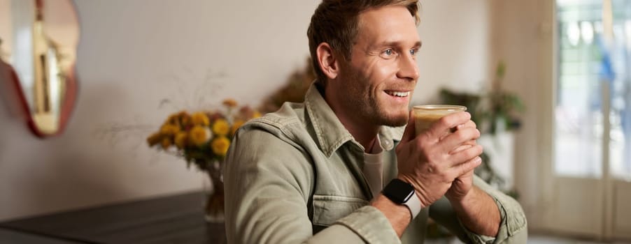 Leisure, people and lifestyle concept. Happy handsome young man in cafe, sitting on chair and smiling, holding glass of coffee, looking aside, relaxing with cup of cappuccino.