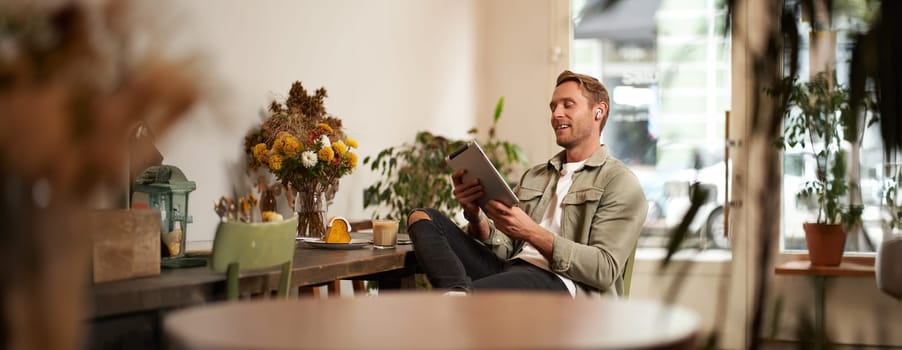 Portrait of handsome smiling young man, sitting in coffee shop, using digital tablet, video chats with someone from a cafe, wearing wireless headphones.