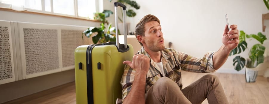 Portrait of happy tourist, man takes selfies before his flight, photos with suitcase, uses smartphone to post pictures on social media.