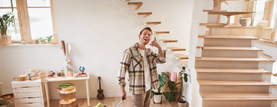 Vertical portrait of happy young man going on holiday, shows mobile phone call hand gesture, walking with suitcase, leaving rented apartment. Travelling and tourism concept