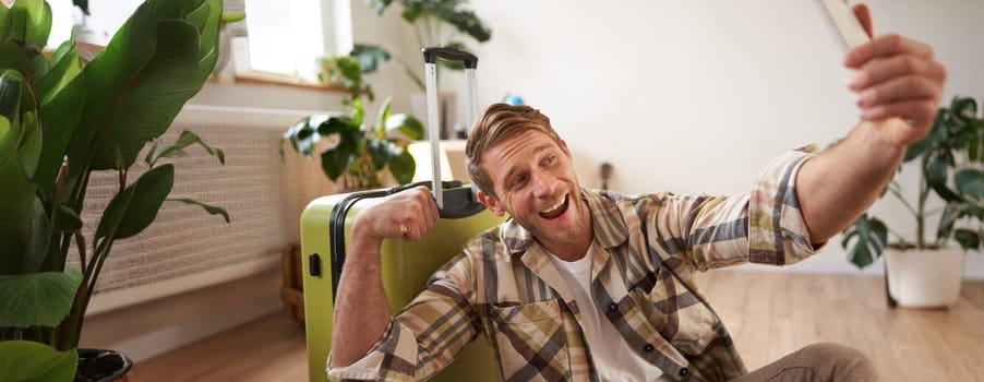 Portrait of smiling handsome guy shows his biceps on photo, takes selfies on smartphone, sits near suitcase, goes on holiday tour.