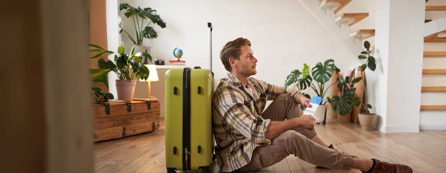 Portrait of smiling handsome tourist, man holding passport and flight tickets, sitting with suitcase. Tourism and travelling concept