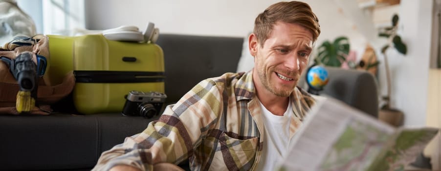 Smiling handsome man with map in hands, packed suitcase, goes on holiday, prepared to go on vacation.