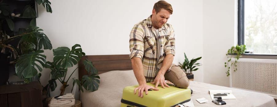 Portrait of young guy, traveller going on holiday, pressing his suitcase, trying to push clothes inside bag, closing his luggage, struggling to pack lots of things for vacation. Tourism concept