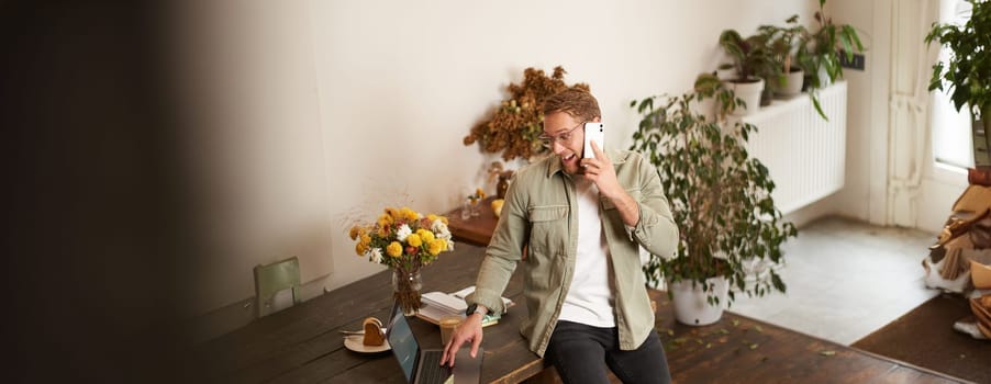 Portrait of happy young manager, man having negotiations, business conversation over the phone, talking on mobile, sitting in office on table with laptop.