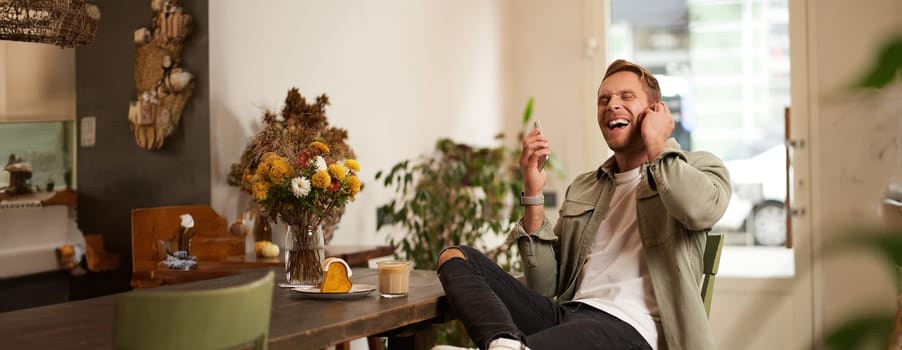 Lifestyle portrait of handsome young man, sitting in cafe, talking to a friend on mobile phone with wireless earphones, laughing and smiling, listening to funny podcast.