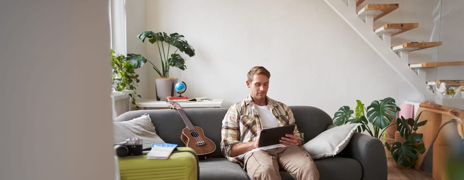 Portrait of young man in living room, sitting with tablet in hands, has suitcase packed and two plane tickets, preparing to go abroad, going on holiday.