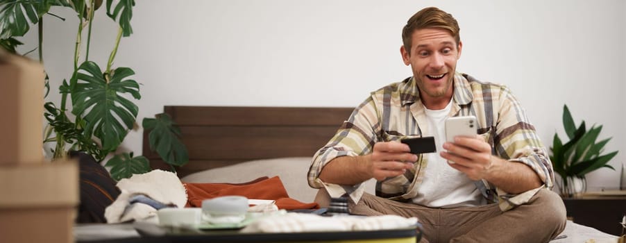 Cheerful male tourist, man packing suitcase, paying for tickets with credit card and smartphone app, celebrating, excited about going on holiday.