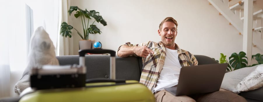 Happy handsome guy, sits with laptop, points finger at screen and laughs, has packed suitcase and camera prepared for holiday, goes on vacation. Tourism and travelling concept