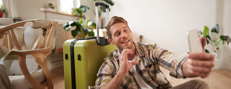 Smiling tourist, young man takes selfie and shows heart sign on phone camera, posing near luggage, packed for holiday tour.
