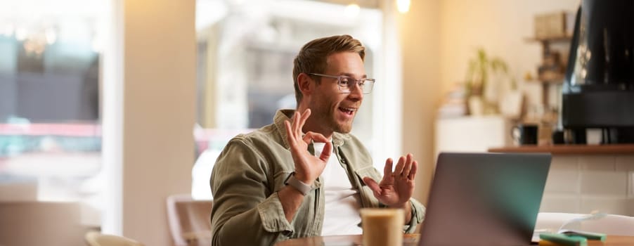 Portrait of young man, teacher, online tutor in glasses, showing okay, ok hand sign, giving lessons remotely from cafe, sitting with laptop, talking to someone on video chat.