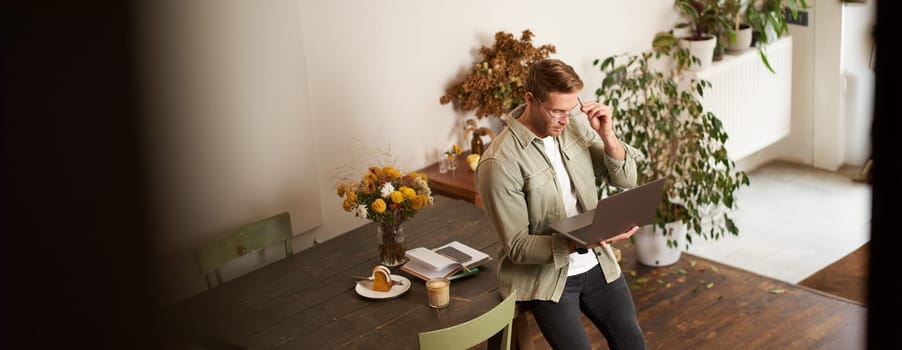 Handsome manager, man in glasses sits on table, holds laptop and types, sends message, works on project, drinks coffee, spends time at co-working space or an office.