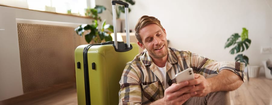 Portrait of handsome smiling man chats on social media, laughs over message on smartphone, looks at mobile phone with happy face, sits near suitcase on floor.