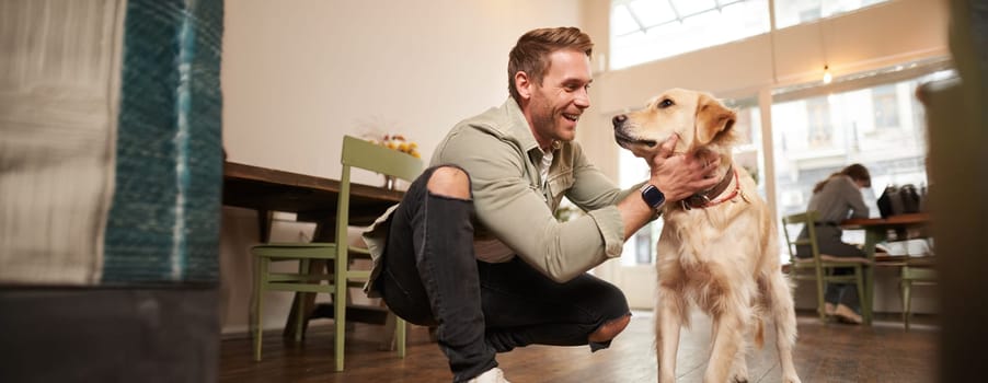 Portrait of happy young man enters coffee shop with his golden retriever. Handsome guy petting dog in pet-friendly cafe.