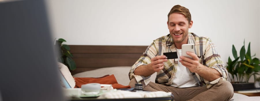 Portrait of young man booking tickets, going on holiday, holding credit card and smartphone, sitting with mobile phone near suitcase filled with clothes, ready for vacation trip.