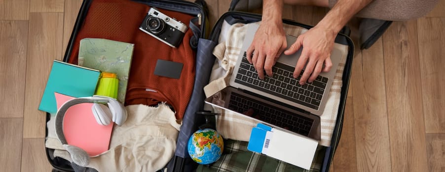 Upper angle view of male hands typing, using laptop on top of the opened suitcase, man packing for holiday, going on vacation, check-in on a flight.