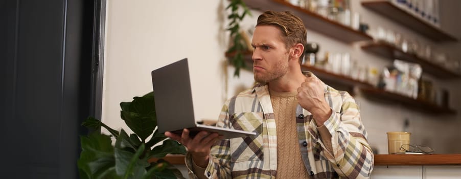 Portrait of angry man in cafe, holding laptop, staring at screen and showing clenched fist during video chat, online meeting.