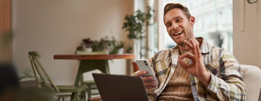 Portrait of handsome man in cafe, sitting with laptop, working remotely, freelancer with phone in hands, shows okay, ok gesture in approval, agree with something, like product.