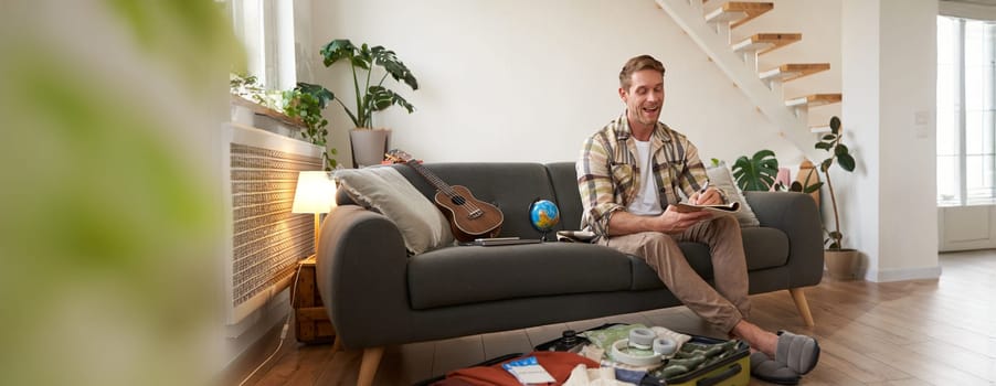 Cheerful handsome man preparing for holiday, going on vacation, writing down, making notes, list of items for travelling, packs suitcase with clothes.