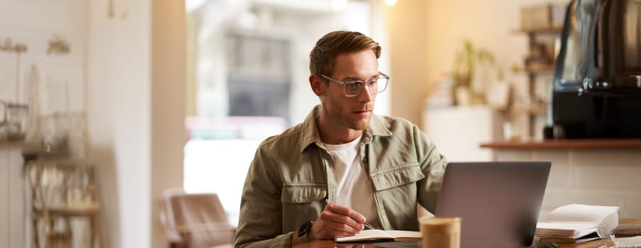 Portrait of modern man in glasses, sitting in quite empty cafe with laptop, writing down notes in notebook, studying online, remote e-learning, passing web course.