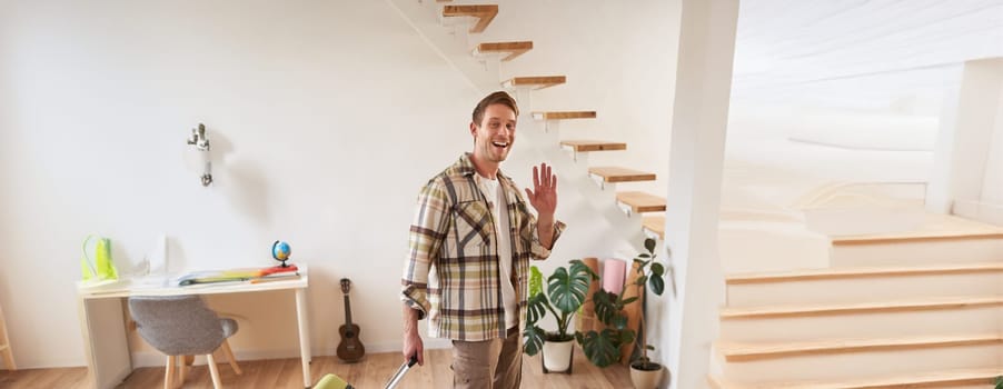 Vertical shot of happy young man with suitcase, checking in the rented apartment, waving hand at camera, walking with luggage indoors.