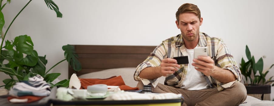 Portrait of man with thoughtful face, sitting on bed with suitcase, paying online with credit card and smartphone app. Copy space
