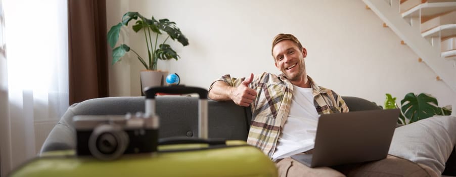 Cheerful man with laptop, tourist sits near suitcase, shows thumb up, books a holiday, goes on vacation. Tourism concept