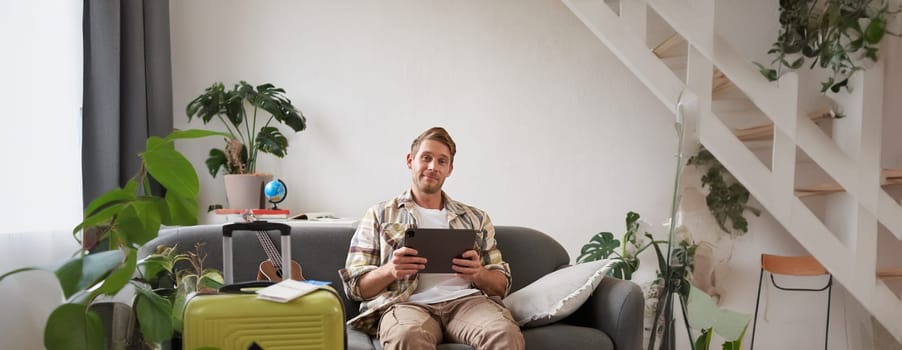 Vertical shot of young man going abroad, packing suitcase for a vacation, holding digital tablet, booking hotel online. Tourism and travel agency concept