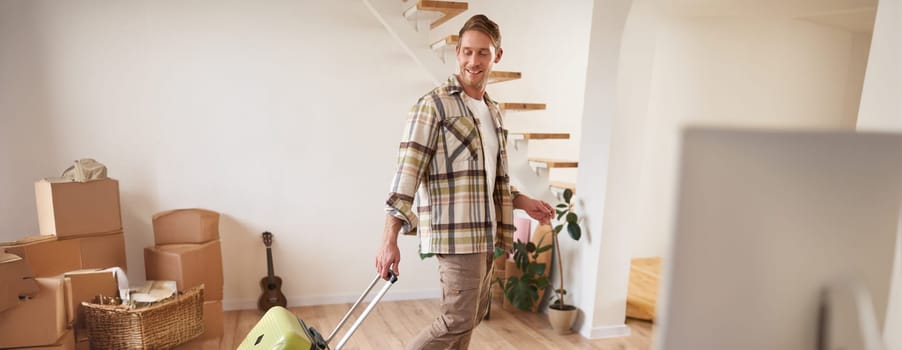 Vertical portrait of happy young man with suitcase, walking indoors, leaving a flat with luggage, going on business trip or vacation. Copy space