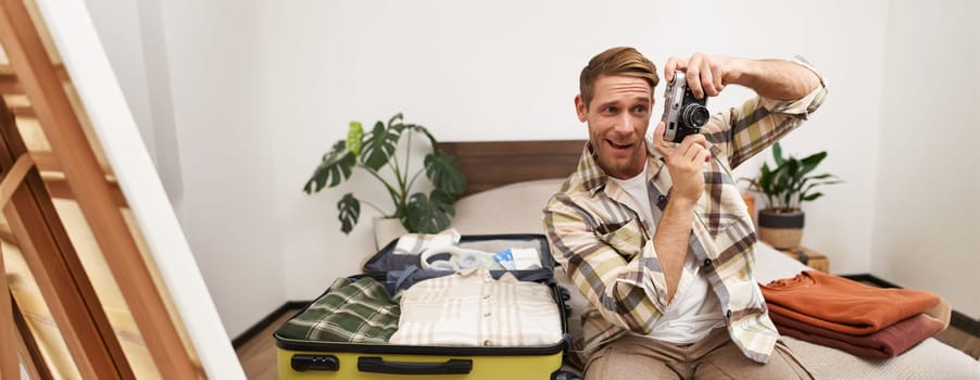 Portrait of handsome man going on vacation, packing suitcase, holding camera, travel photographer going on holiday.