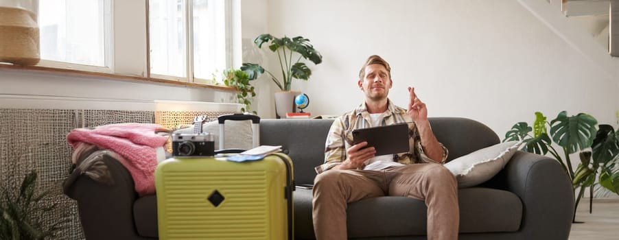 Portrait of man waiting for his holiday tour confirmation, cross fingers, hopes for something, holds tablet, has suitcase packed and ready for holiday.