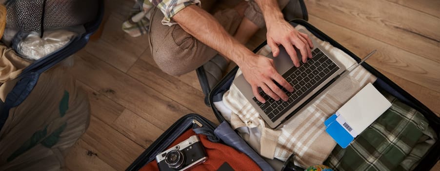 Close up shot of male hands, tourist using laptop, sitting on floor with opened suitcase, packed luggage for holiday, has two flight tickets, camera and clothes.