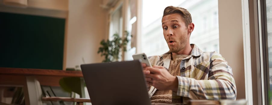 Image of man in cafe, coffee shop visitor sitting in chair with laptop and smartphone, looking surprised and excited at mobile phone screen, amazed by big news, promo offer on app.