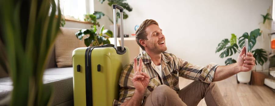 Portrait of smiling handsome man, shows peace sign, takes selfies on smartphone, sits with suitcase, goes on holiday. Tourism and travel agency concept