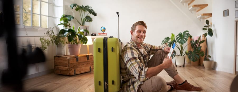 Portrait of handsome young man, tourist sitting with his suitcase, holding passport and flight tickets, going on holiday.