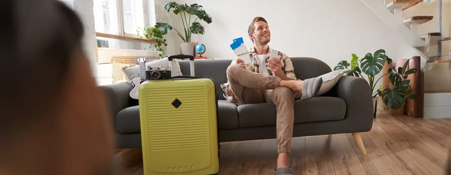 Handsome young man sits on sofa with suitcase, holds smartphone, books hostel or holiday vacation, chats on mobile phone. Tourism and travelling concept