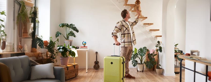 Portrait of happy traveller, young man going on vacation, holding his suitcase, petting a cat on stairs. Tourism and people lifestyle concept